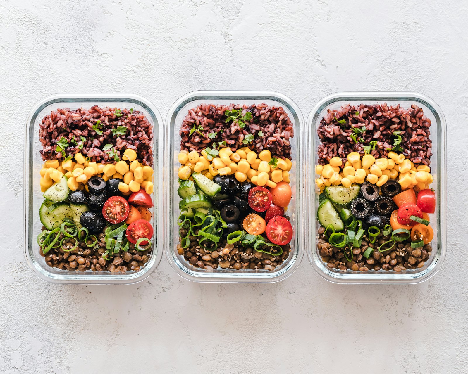 Trois boites de repas en verre garnies de riz, mais, concombre, tomates, olives et lentilles, pour transformer des restes en repas equilibre.